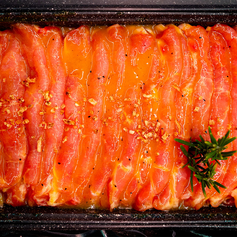 Artigianalità Affettati di carne cruda marinati e conditi in una teglia pronti per la cottura, guarniti con un rametto di rosmarino